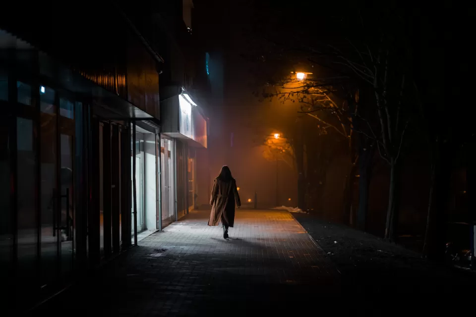 Silhouette of woman in long coat walking on darkened city street alone at night, passing brightly lit sotrefront.